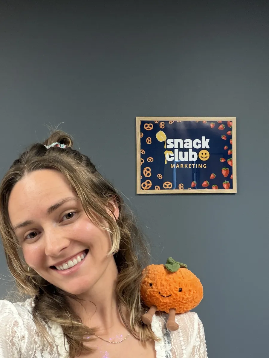 Bre smiling with the plush orange on her shoulder in front of a Snack Club sign