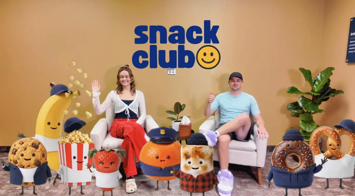 Joe and Bre sitting in the office surrounded by the whole Snack Club mascot crew